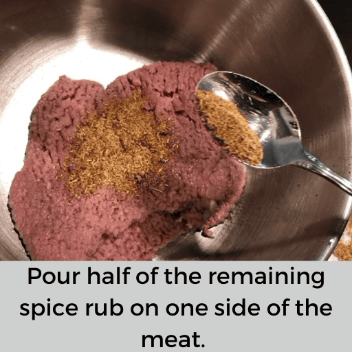 A spoonful of the gluten free country fried steak spice rub is being dumped on the beef cubed steak in the metal bowl.