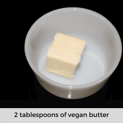 two tablespoons of vegan butter in a white dish sitting on a black stovetop