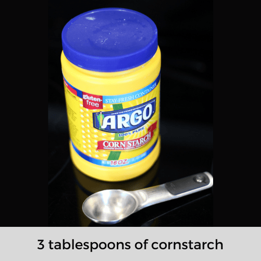 A container of cornstarch on a black surface with a 1 tablespoon sitting in front of it
