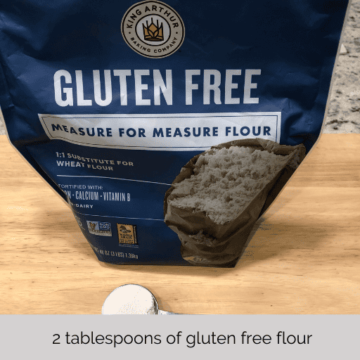 a tablespoon of flour in a measuring spoon sitting on a cutting board in front of a bag of gluten free flour