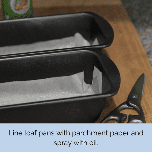 2 loaf pans lined with parchment paper sitting on a cutting board. A pair of scissors is to their right and a can of cooking spray is sitting behind them. 