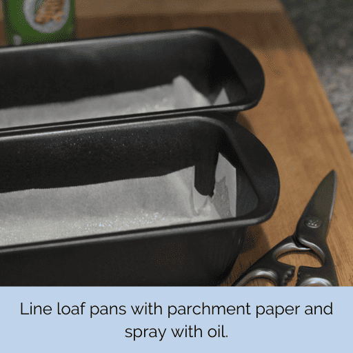2 loaf pans lined with parchment paper sitting on a cutting board. A pair of scissors is to their right and a can of cooking spray is sitting behind them.