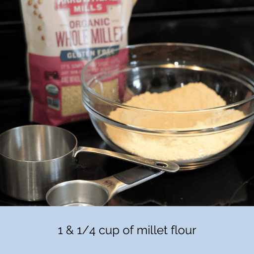 A glass bowl of freshly ground millet flour sitting on a black stovetop. Beside the bowl are two measuring cups. Behind the bowl is a bag of millet grains.