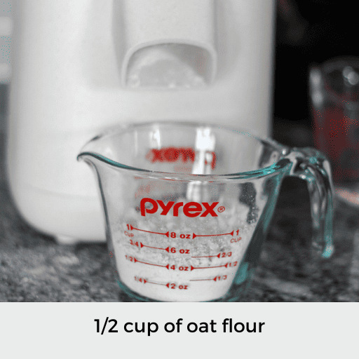 glass measuring cup filled to the 1/2 cup measuring line with oat flour. The measuring cup is in front of a grain mill.
