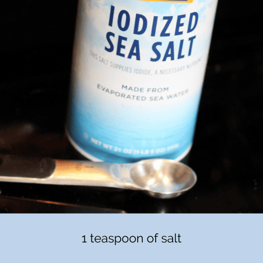 A 1 tsp measuring spoon in front of a container of sea salt on a black stovetop