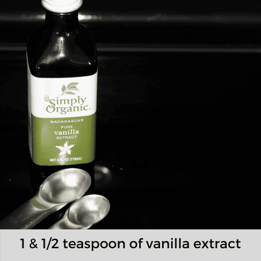 a glass bottle of simply organic vanilla extract with a teaspoon and a half teaspoon sitting in front of it on a black stovetop. 