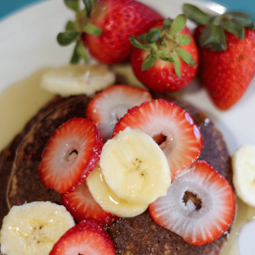 gluten and dairy free pancakes topped with maple syrup and sliced bananas and strawberries. There are three whole strawberries in a group on the plate 