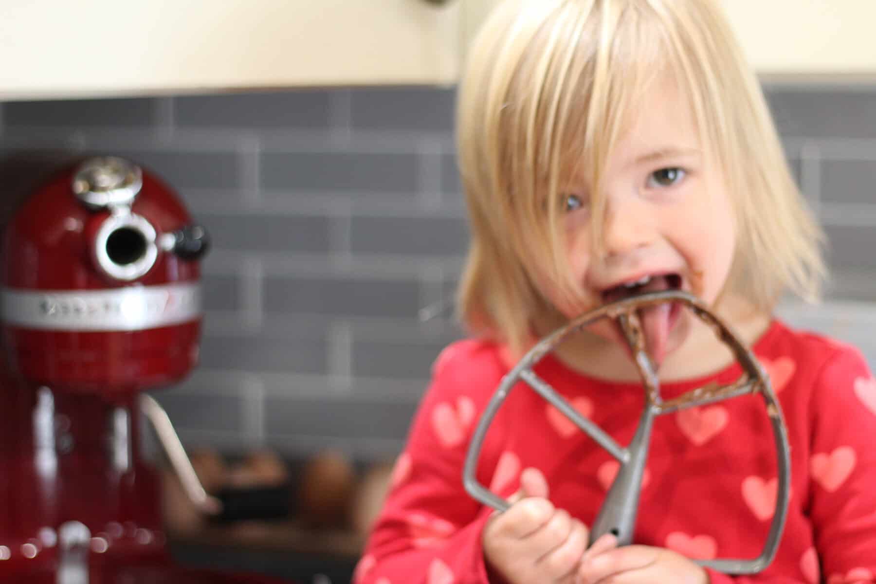 little girl licking the dairy free chocolate brownie icing off the paddle attachment 