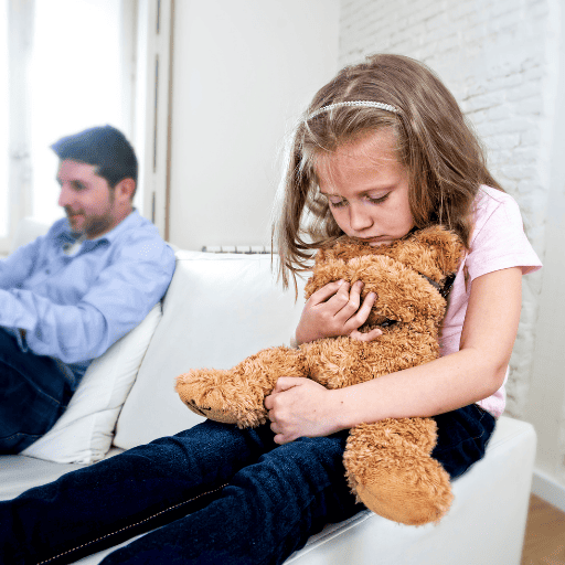 little girl looking sad holding a teddy bear while sitting on a arm of a couch. Her father is sitting happily on the couch ignoring her