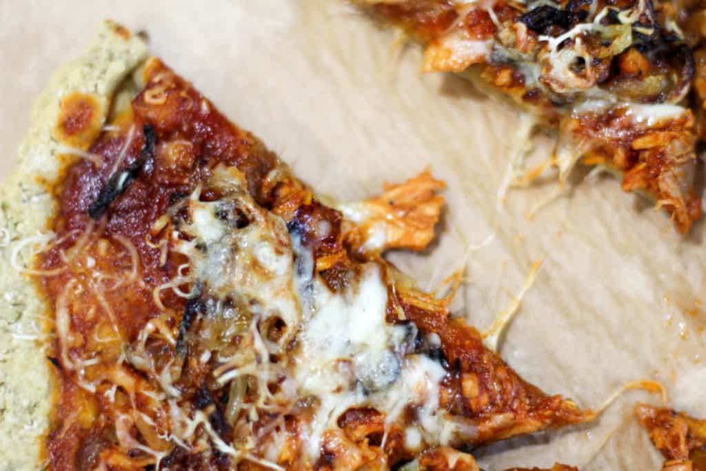 a close up of a slice of gluten and dairy free pizza crust topped with BBQ sauce, BBQ chicken, fried onions, and cheese. 