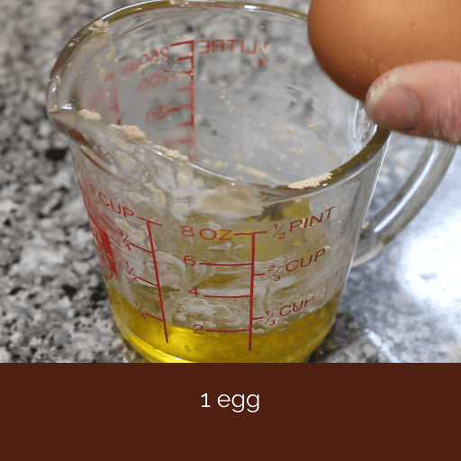 egg being cracked on the edge of a glass measuring cup filled with olive oil. 