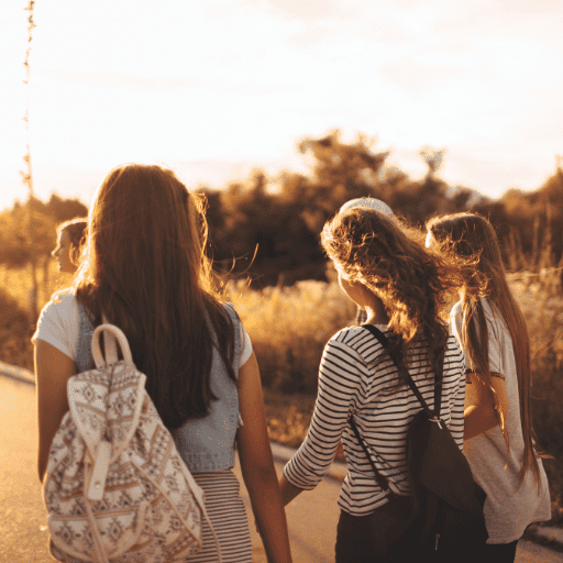three happy teenage girls holding hands walking at dusk. Two are wearing backpacks. There is gold lighting all around them. 