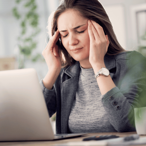 young adult woman sitting in front of a computer holding her temples because of a headache.