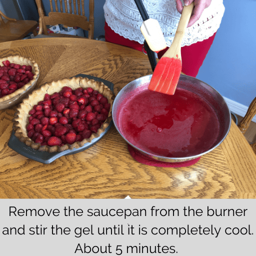 saucepan of gel sitting beside the pie crust filled with strawberries on a wooden table.