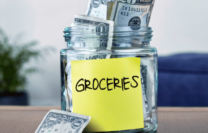 glass jar of money with a yellow sticky note that says groceries on it
