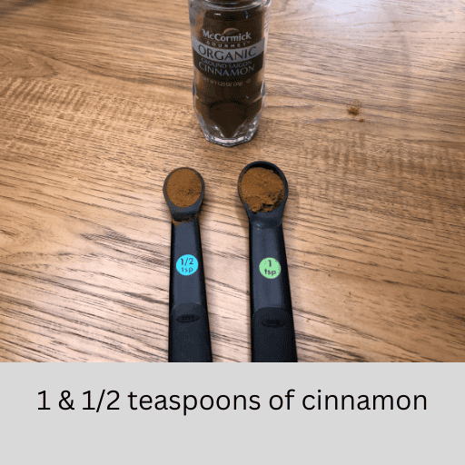 one and a half teaspoons of cinnamon measured out sitting in black measuring spoons of a wooden countertop in front of a jar of organic cinnamon