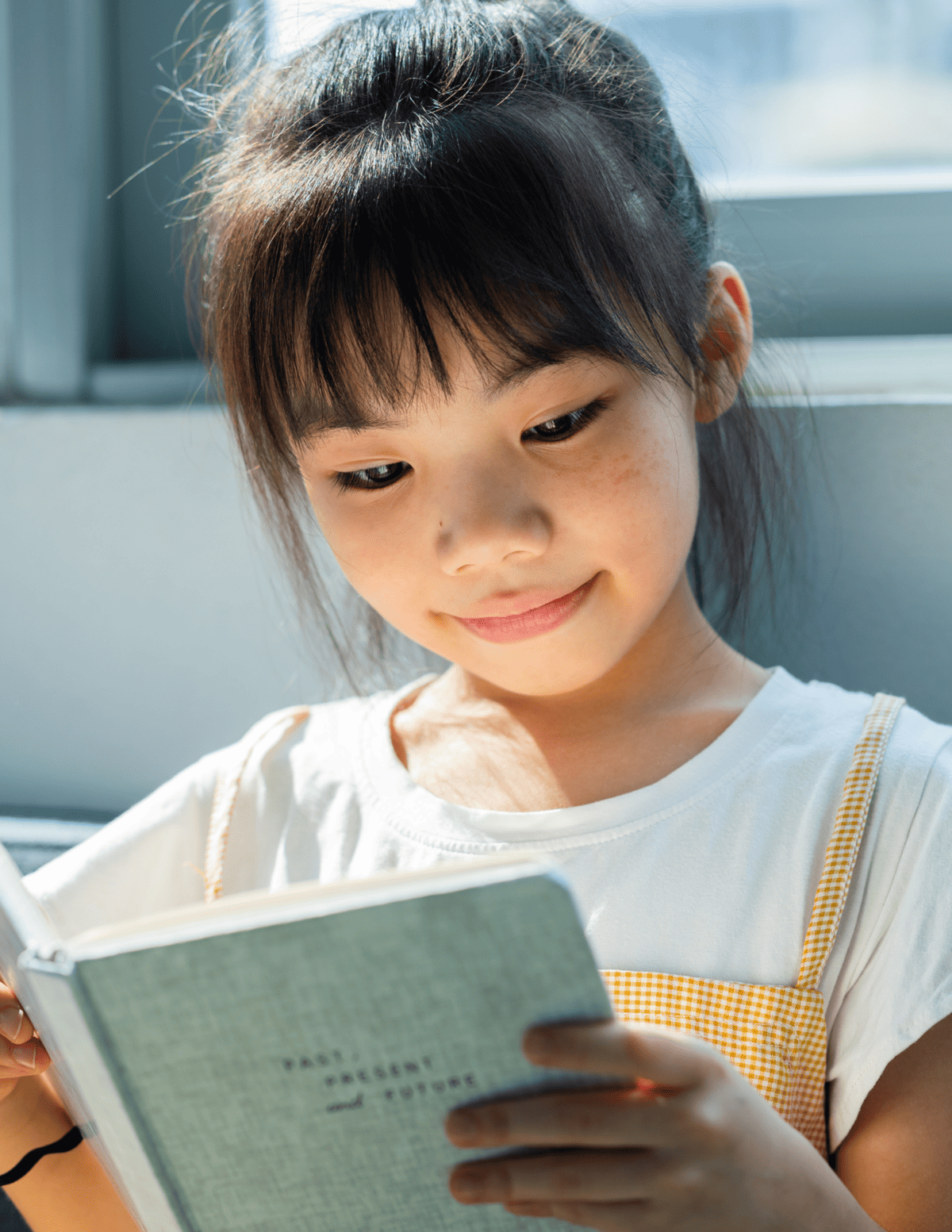 A little girl with black hair smiling, sitting on a couch holding a booklet and reading