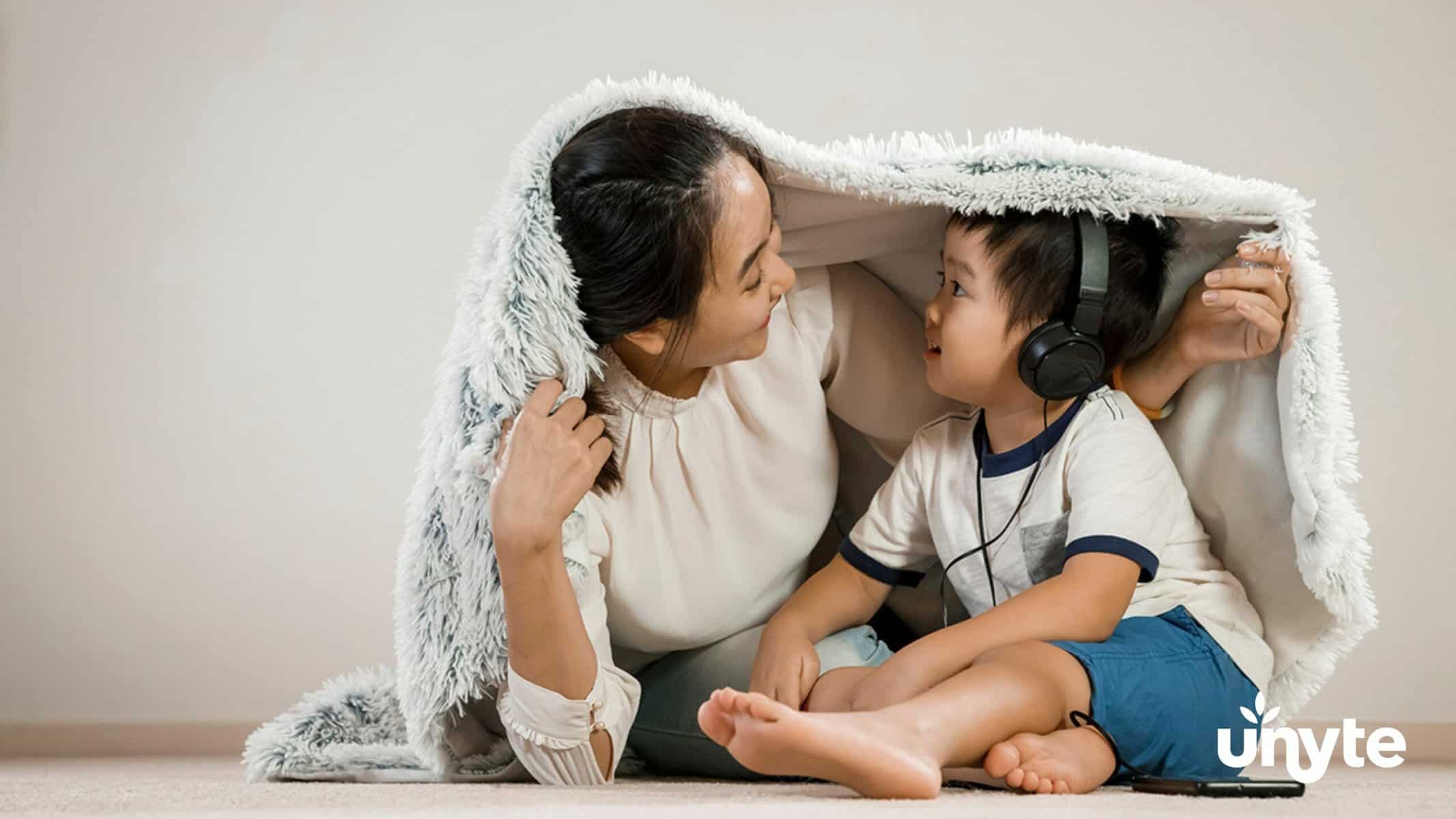 Preschooler wearing blue shorts, white T-shirt, and black headphones looking at mom. They are both under a blanket.